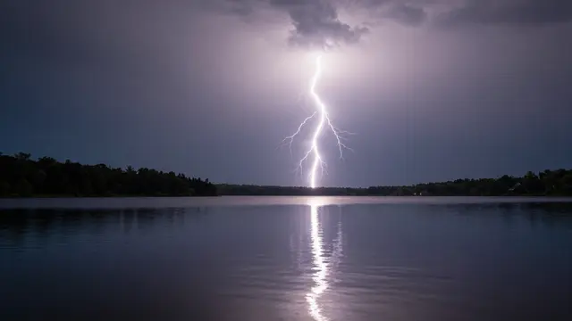Bright lightning bolt strikes above a still lake, its glowing reflection stretching across the calm water under a stormy