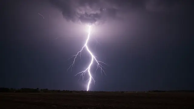 Bright jagged lightning bolt illuminates dark storm clouds above a flat night landscape