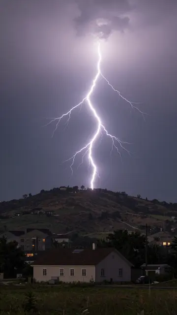 Bright jagged lightning bolt strikes a distant hilltop above a residential house during a night thunderstorm