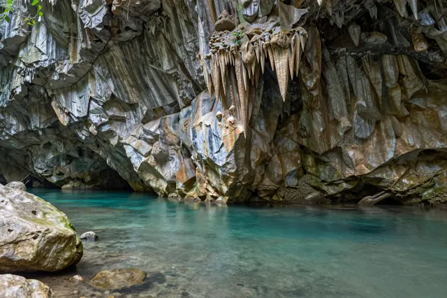 A clear turquoise underground river flows beneath jagged stalactite formations inside a limestone cave