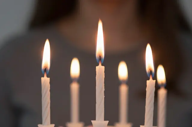 Sharp close-up of lit twisted birthday candle flames, with an out-of-focus person in the background preparing to blow them