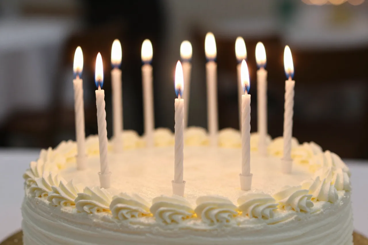 A frosted white birthday cake with multiple lit plain candles sits against a blurred party background.
