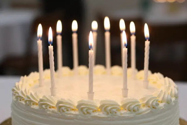 A frosted white birthday cake with multiple lit plain candles sits against a blurred party background.