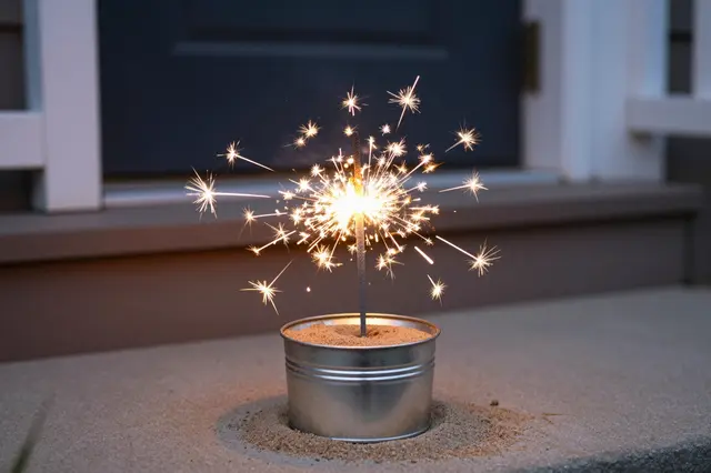 A burning sparkler stands in a small metal sand pail on a home porch step at dusk.