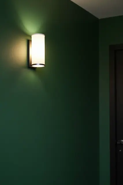 A lit cylindrical wall sconce casts warm light on a deep green interior hallway wall