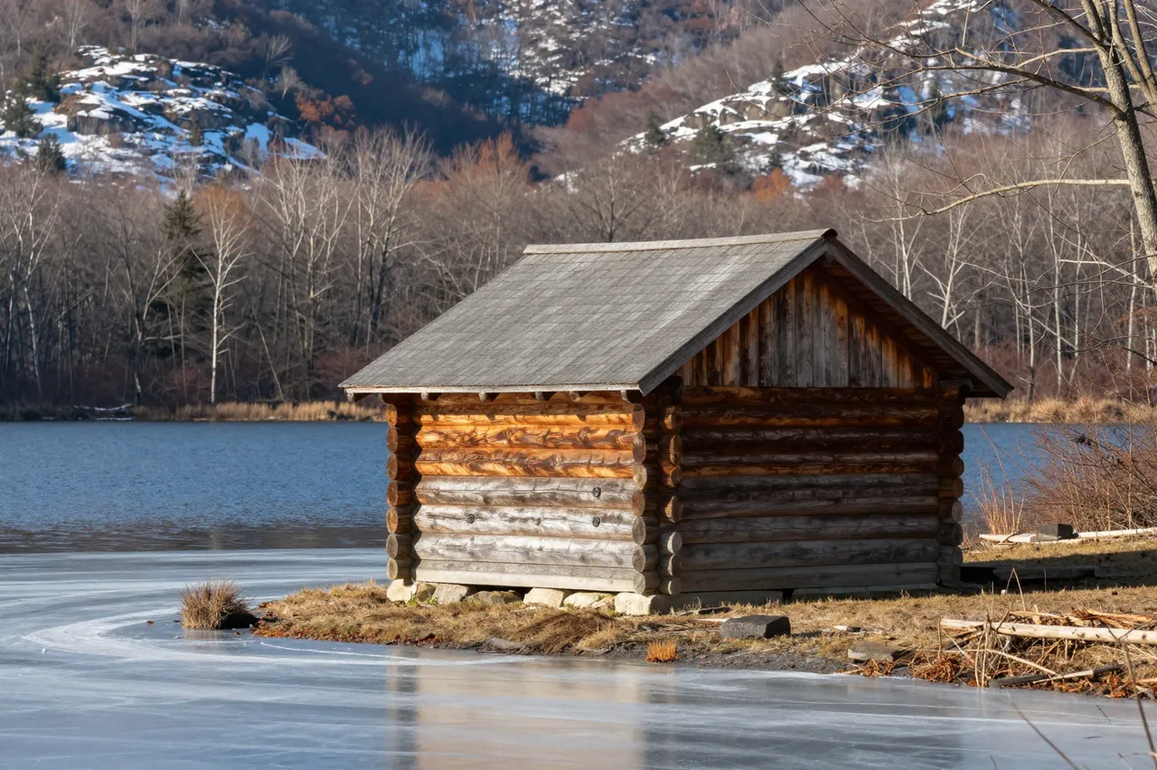 Small log cabin sits on a lakeshore with thin shore ice and snow-dusted distant hills in late autumn