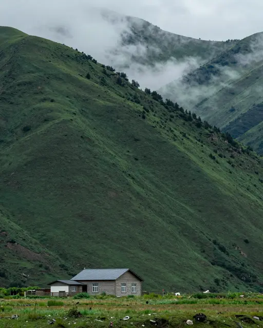 A small stone farmhouse sits at the foot of green mountains partially shrouded in drifting cloud