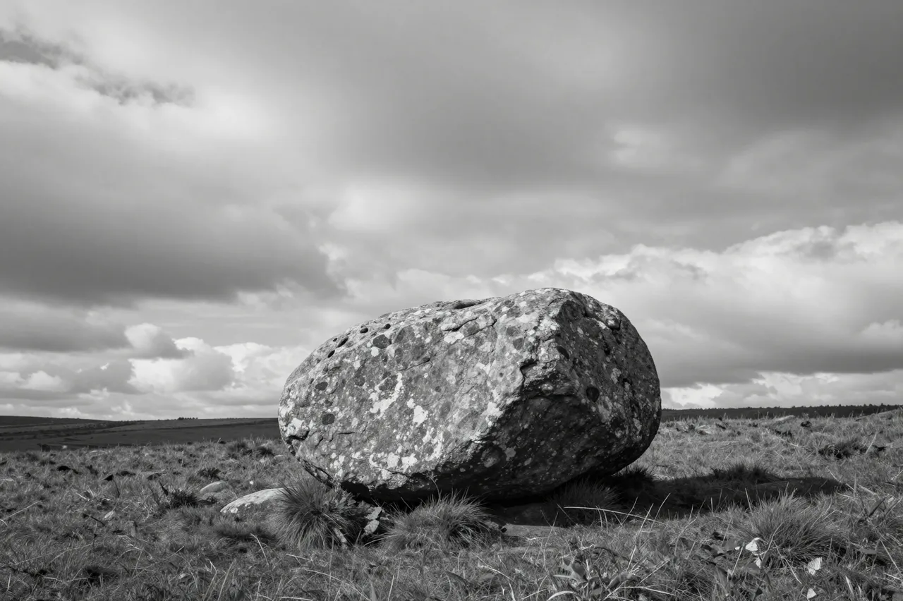 A large weathered boulder sits alone on a grassy moor beneath a dramatic cloudy sky in black and white.