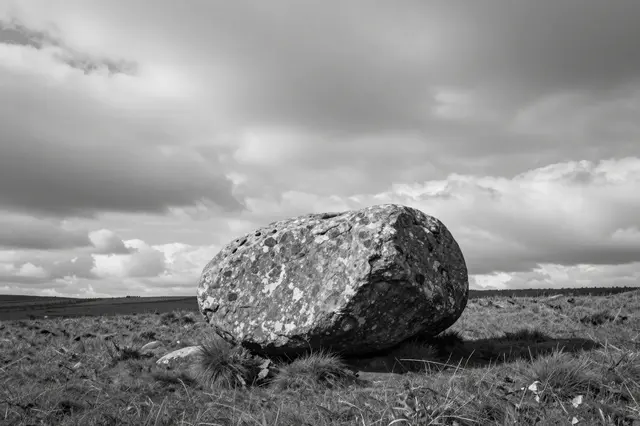 A large weathered boulder sits alone on a grassy moor beneath a dramatic cloudy sky in black and white.