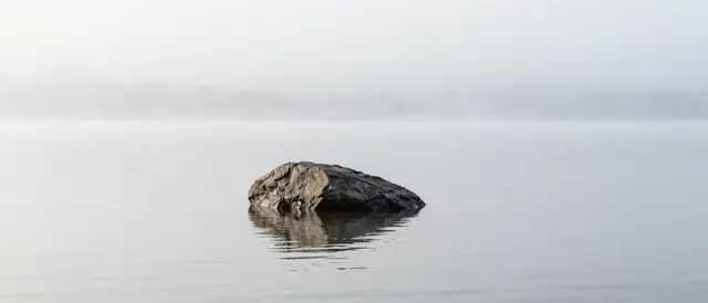 A single weathered rock sits in perfectly still calm lake water under thick white fog