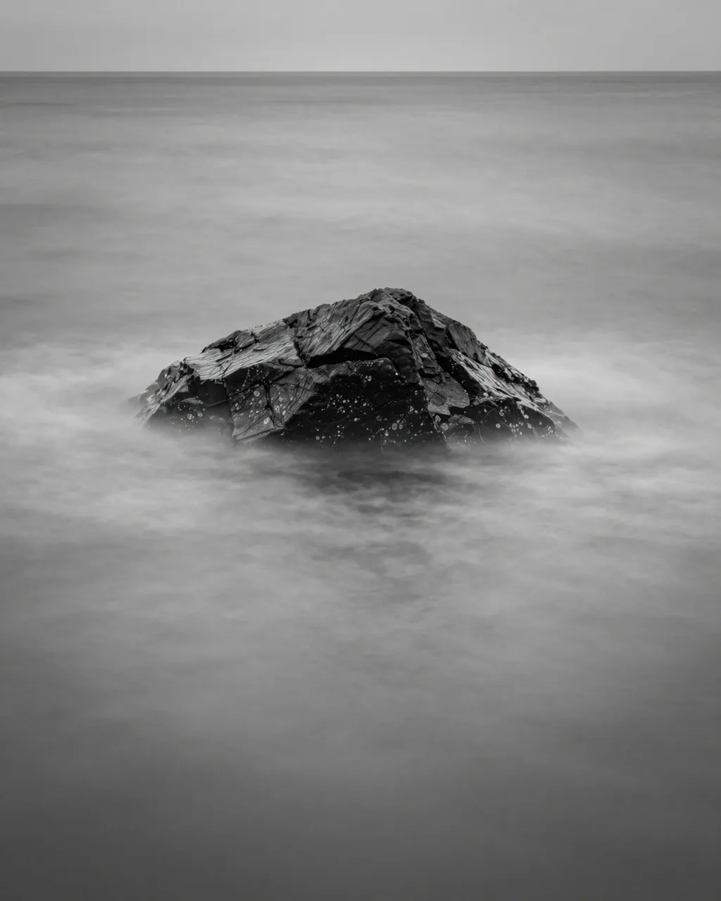 Black and white long exposure view of a lone jagged rock surrounded by silky smoothed sea water
