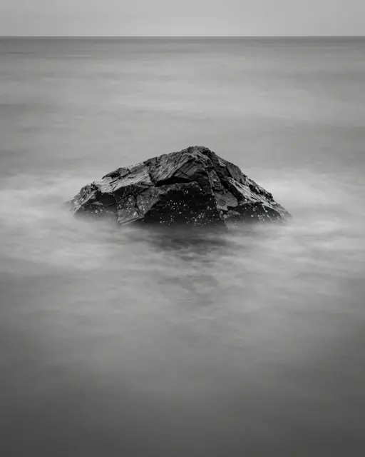 Black and white long exposure view of a lone jagged rock surrounded by silky smoothed sea water