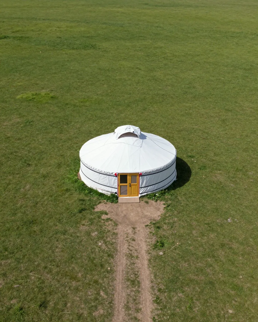 Aerial view of a white yurt with a wooden door on an open grassy steppe, with a dirt track leading to its entrance