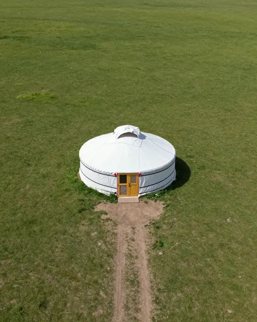 Aerial view of a white yurt with a wooden door on an open grassy steppe, with a dirt track leading to its entrance