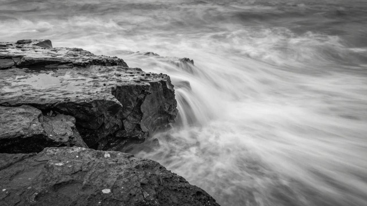 Black and white long exposure of misty ocean waves flowing over a rocky shore shelf