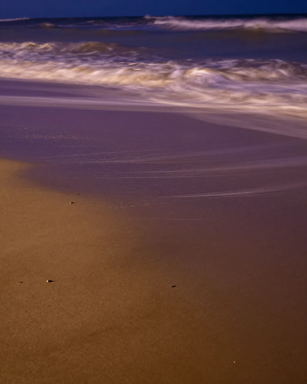 Rippled long-exposure ocean waves wash over wet purple and gold beach sand at night