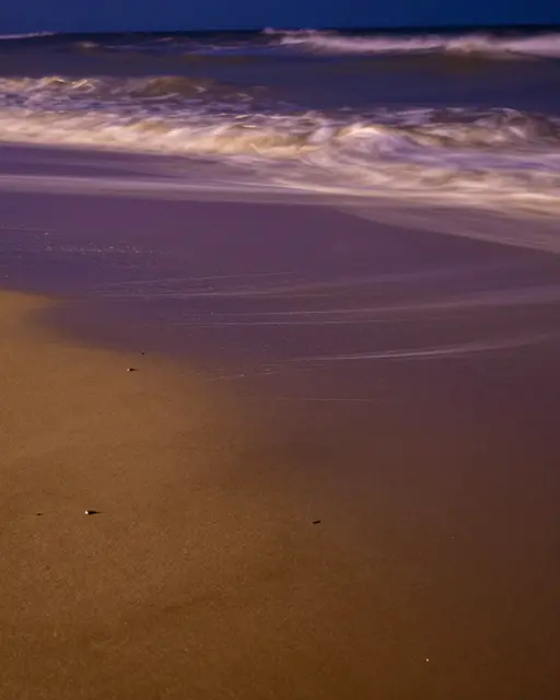 Rippled long-exposure ocean waves wash over wet purple and gold beach sand at night