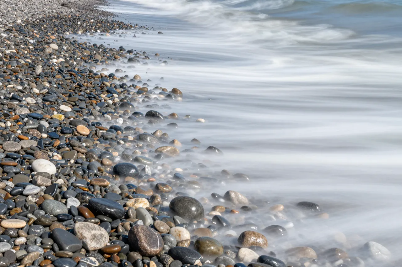 Smooth long exposure sea waves wash over multicolored smooth beach pebbles along a shoreline