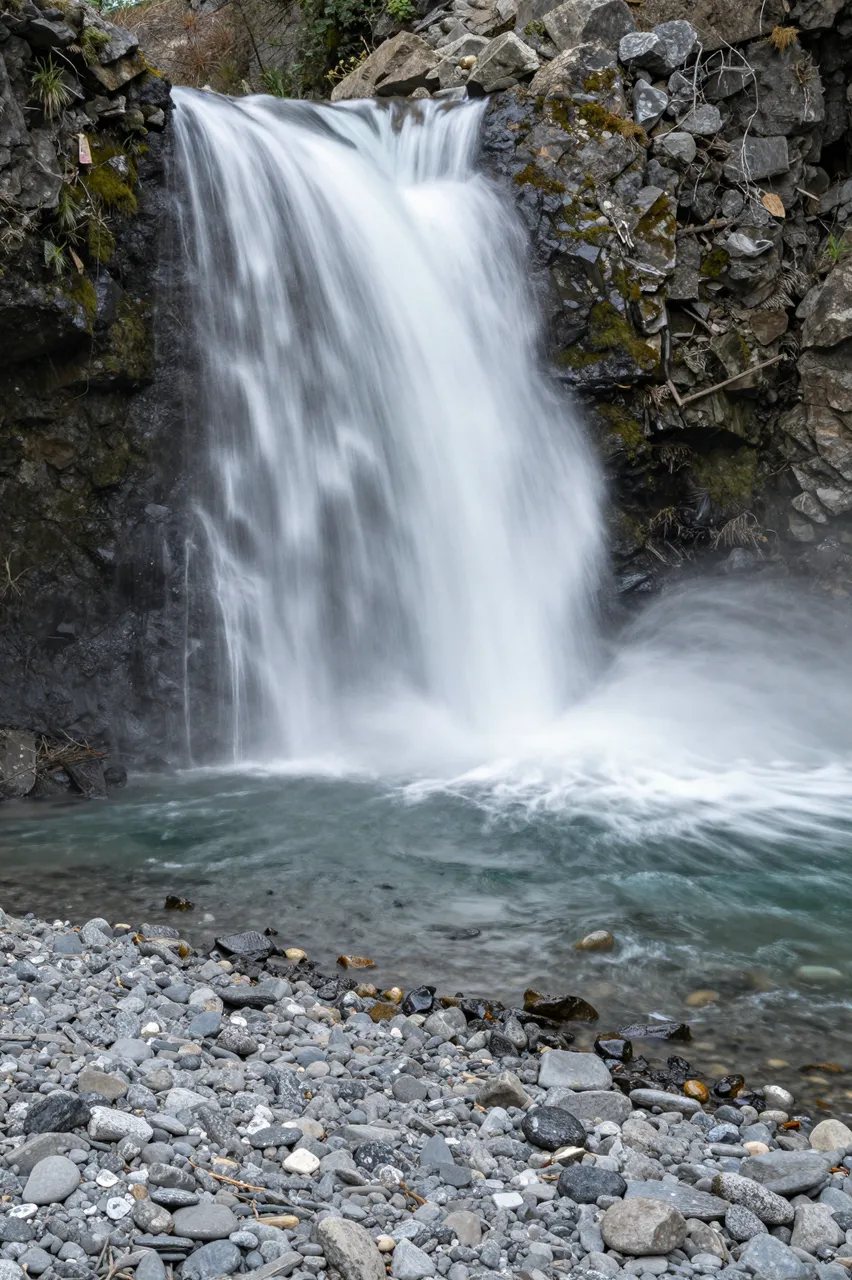 Silky blurred waterfall flows into clear water beside a gray pebble shore framed by mossy rocks