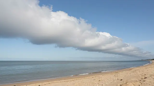A long horizontal roll cloud stretches across a clear blue sky above a quiet sandy beach and calm sea.