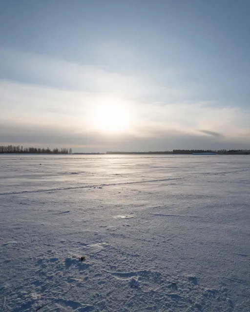 Pale low winter sun hangs over a vast snow-covered frozen plain under a soft blue sky