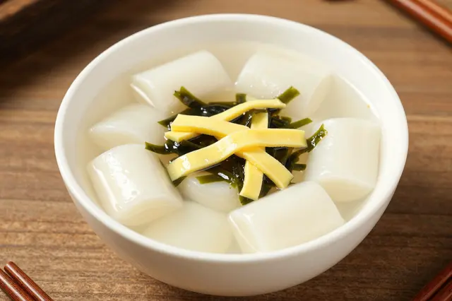 A white bowl of rice cake soup topped with egg ribbons and seaweed rests on a wooden table