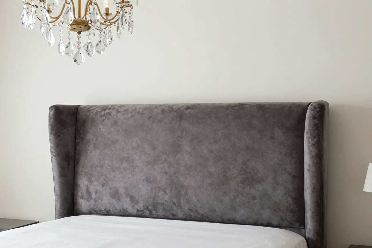 Gray velvet wingback headboard sits below a crystal and brass chandelier in a bright minimalist bedroom.