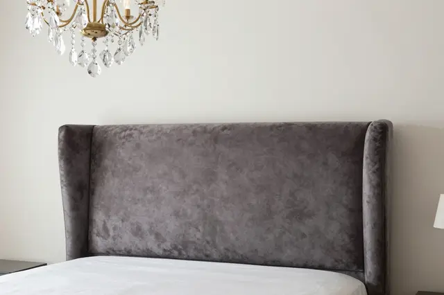 Gray velvet wingback headboard sits below a crystal and brass chandelier in a bright minimalist bedroom.