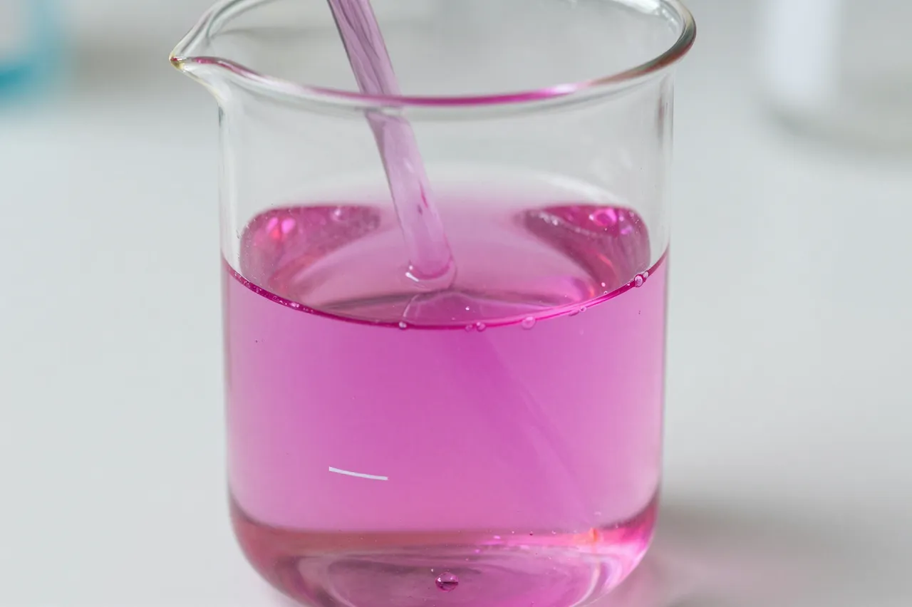 A glass beaker holds vibrant magenta liquid with an inserted glass stirring rod on a lab bench.