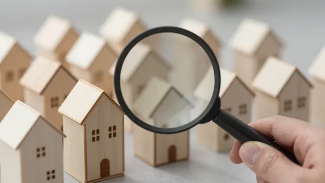 A hand holds a magnifying glass over a row of small wooden house models for a home search concept