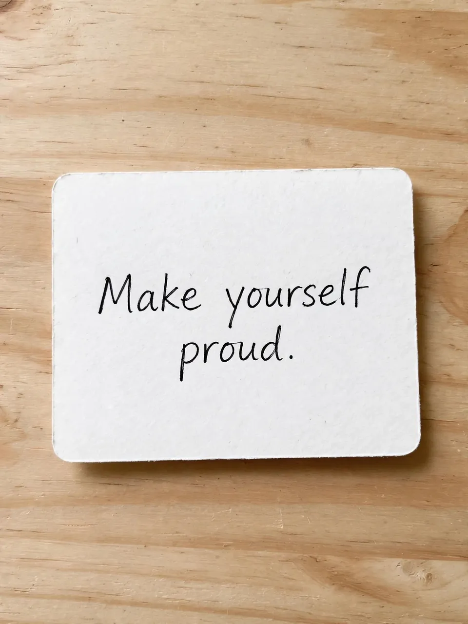 Handwritten note reading "Make yourself proud." rests on a light wooden desk