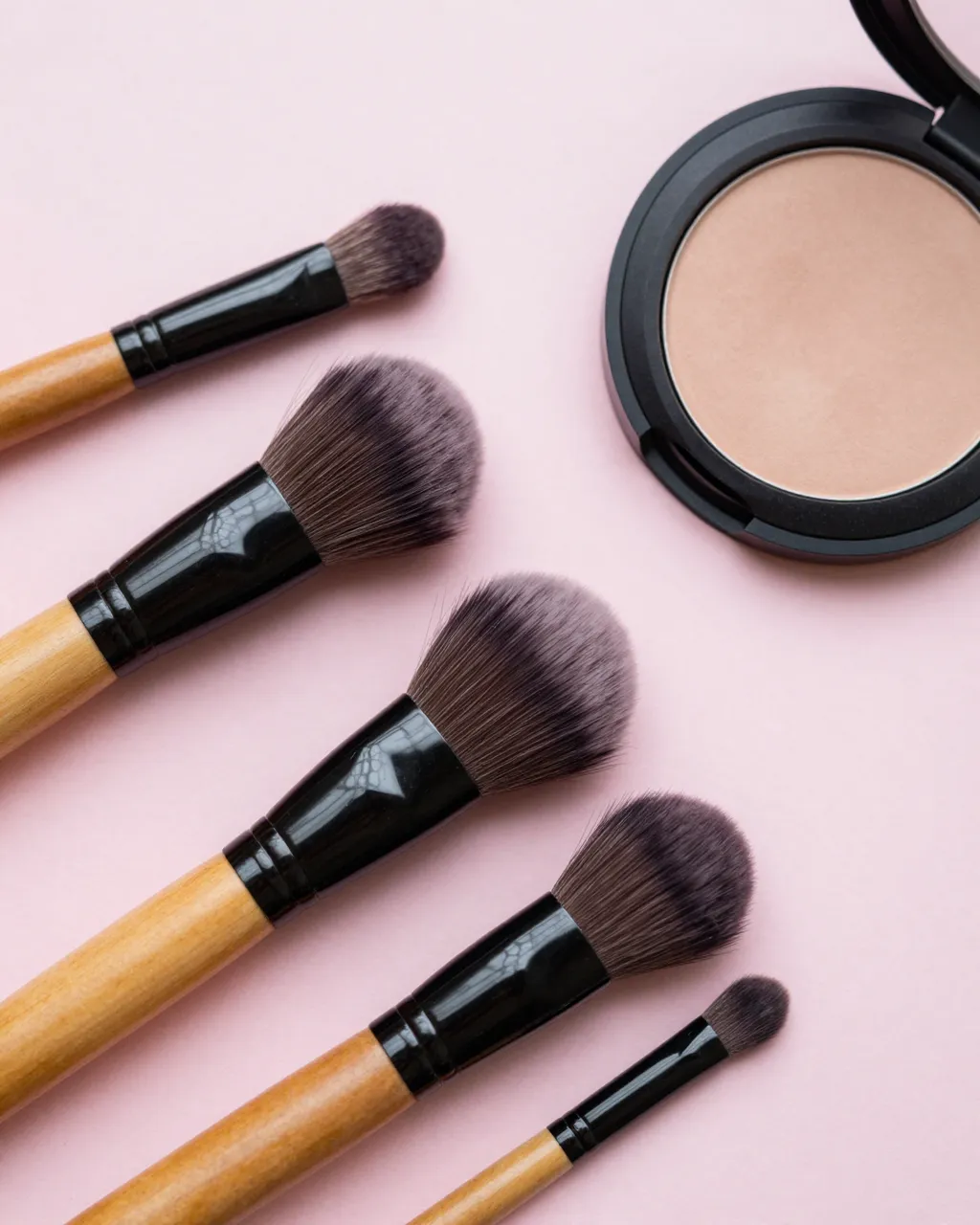 Top-down flat lay of wooden-handled makeup brushes next to an open powder compact on a pale pink background
