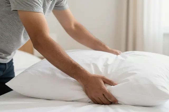 A man adjusts a white pillow on a neatly made bed in a sunlit bedroom