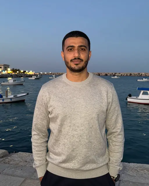 Middle Eastern man in a gray sweater stands on a stone harbor pier at dusk with small boats on the calm water behind him