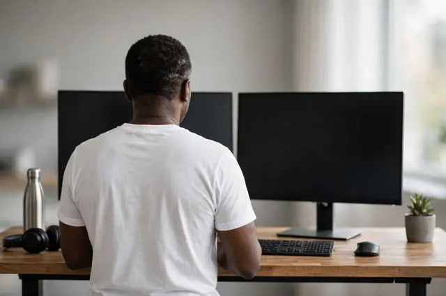 Back view of a Black man in a plain white shirt working at a standing desk with two blank monitors