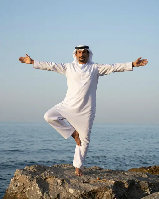 Middle Eastern man in traditional clothing holds a tree pose balancing on one leg on a seaside rock at dawn