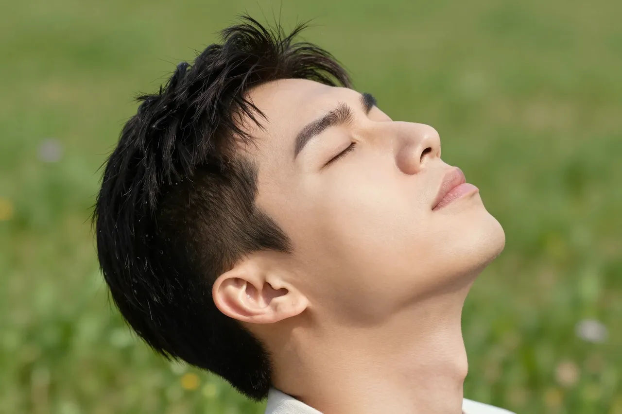A close-up side view of a young adult man with closed eyes tilting his head back in a sunlit grassy meadow.