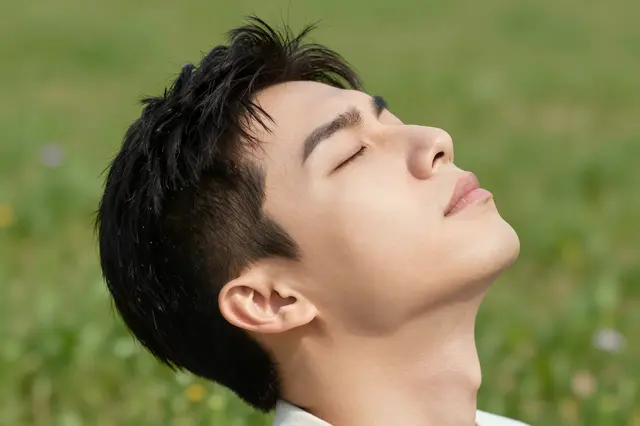 A close-up side view of a young adult man with closed eyes tilting his head back in a sunlit grassy meadow.