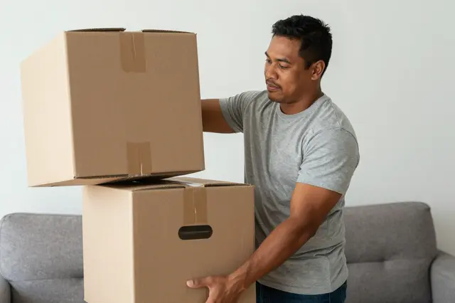 Pacific Islander man in a gray t-shirt holding stacked cardboard moving boxes near a gray sofa