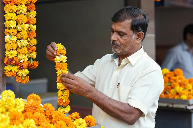 A South Asian man in his fifties examines a marigold garland at a flower market stall.