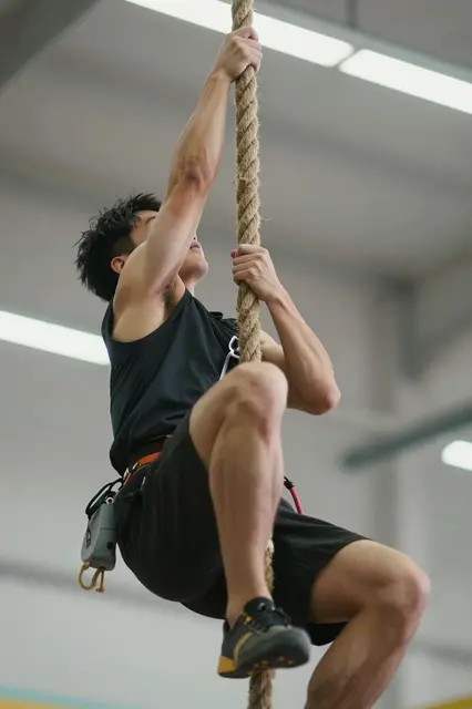 Low angle view of a fit man gripping a thick rope while climbing inside a bright indoor gym.