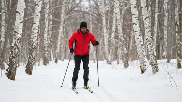 A Black man in a red jacket cross-country skis along a snowy path through a winter birch forest