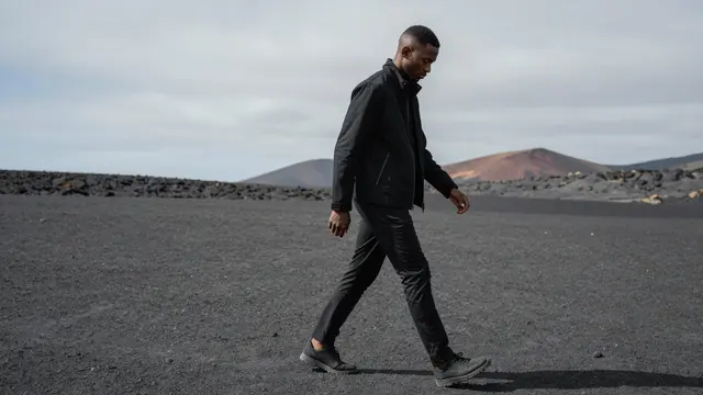 A Black man in a black jacket walks slowly across a dark volcanic sand plain under a gray overcast sky