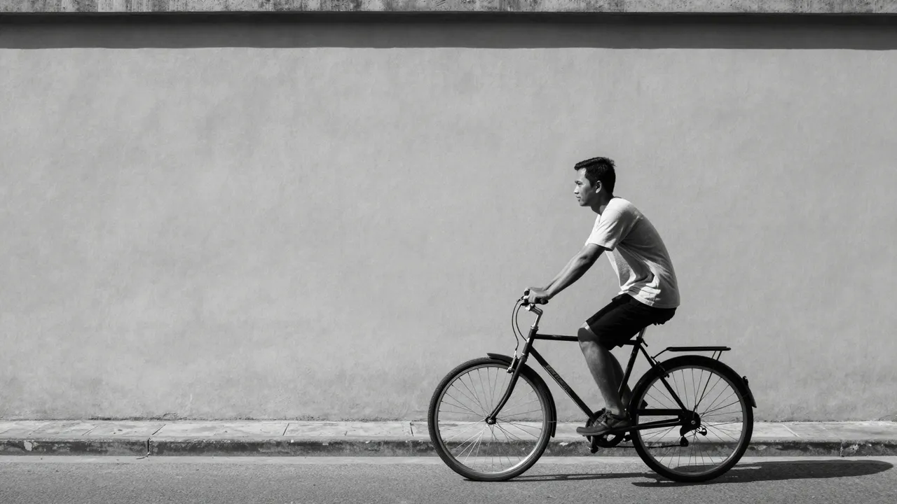 Black and white shot of a Southeast Asian man riding a bicycle along a street beside a plain long wall