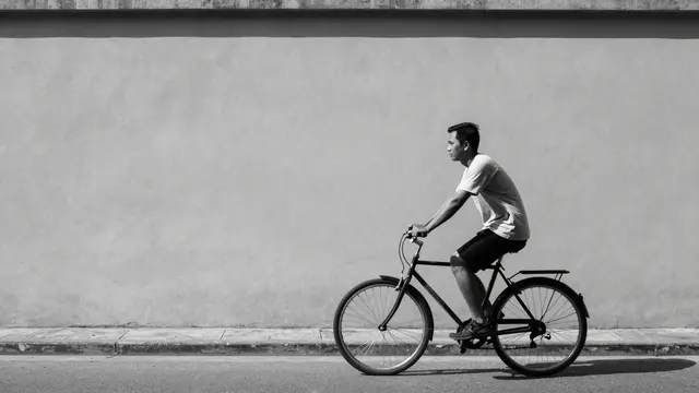 Black and white shot of a Southeast Asian man riding a bicycle along a street beside a plain long wall