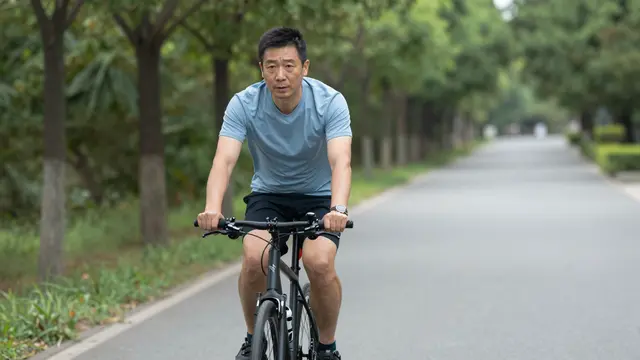 Middle aged East Asian man rides a bicycle along a paved tree lined park path
