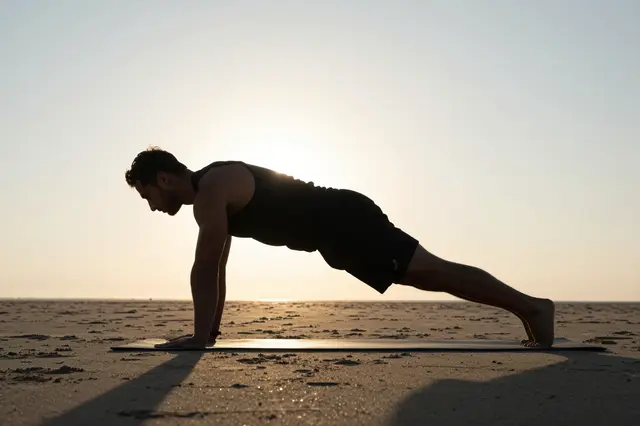 Silhouetted adult man holds a plank exercise pose on a yoga mat atop sand at sunrise