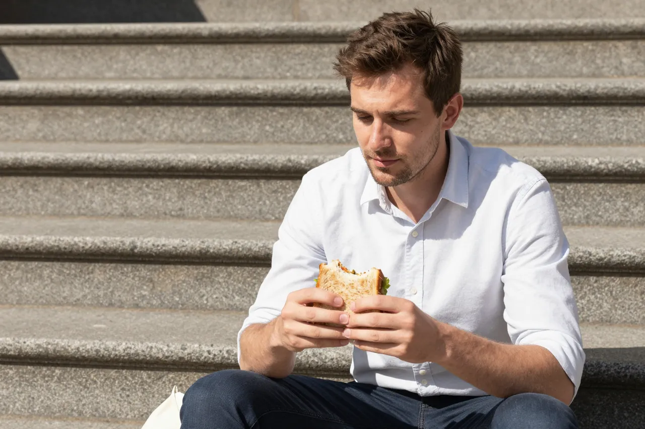 A man in a plain shirt sits on sunny stone steps holding a sandwich during his lunch break