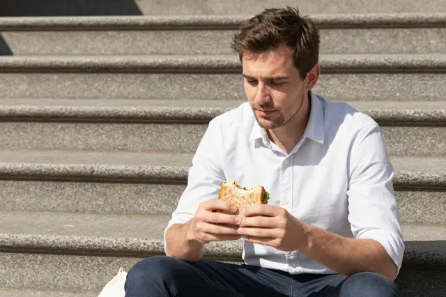 A man in a plain shirt sits on sunny stone steps holding a sandwich during his lunch break