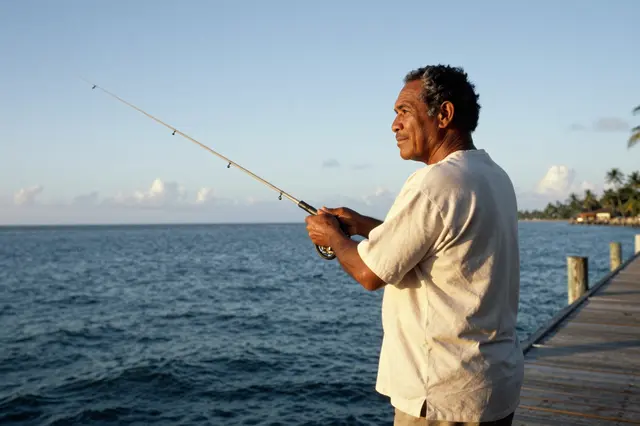 Senior Pacific Islander man holds a fishing rod while standing on a wooden pier overlooking the ocean at sunset